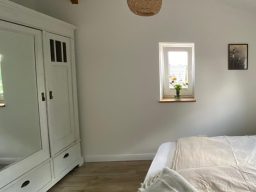 Schlafzimmer mit Bett, weißem Kleiderschrank und Fenster mit Blumen.