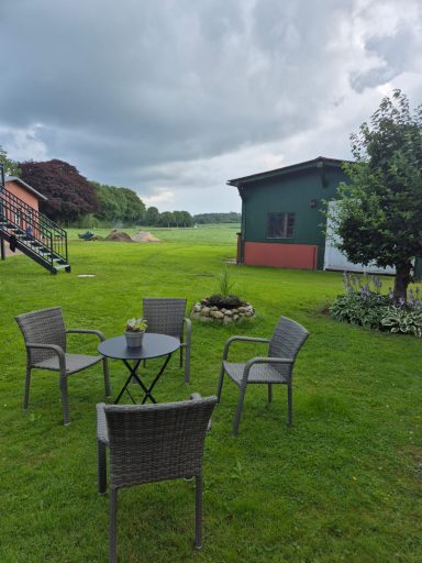 Vier Stühle und ein Tisch im Garten, umgeben von Rasen und Bäumen, mit bewölktem Himmel.