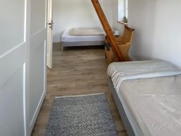 Heller Raum mit zwei Betten und einem Teppich, Holztreppe im Hintergrund.