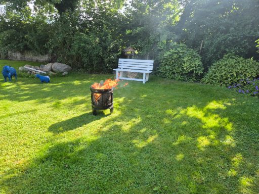 Feuerstelle im Garten mit grünem Rasen und einer weißen Bank im Hintergrund.