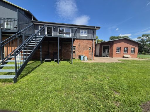 Zweistöckiges Gebäude mit Terrasse und Nebengebäude, umgeben von Wiese.