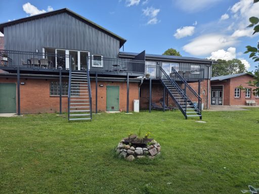 Zwei zweigeschossige Gebäude mit Treppe und Feuerstelle im Vordergrund, umgeben von Rasen.