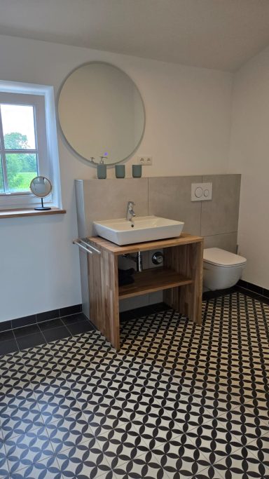 Modernes Badezimmer mit Holzwaschtisch, runder Spiegel und schwarzem Fliesenboden.