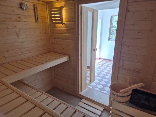 Holzsauna mit zwei Bänken, offener Tür zum angrenzenden Raum und einem Eimer.