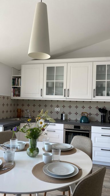 Helle Küche mit weißem Mobiliar, gedecktem Tisch und Blumenstrauß in einer Vase.