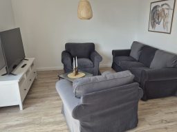 Wohnzimmer mit zwei grauen Sesseln, einer hellen Couch, TV und neutraler Dekoration.