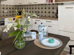 Tischgedeck mit blumigem Arrangement, Teekanne und Tellern in einer modernen Küche.