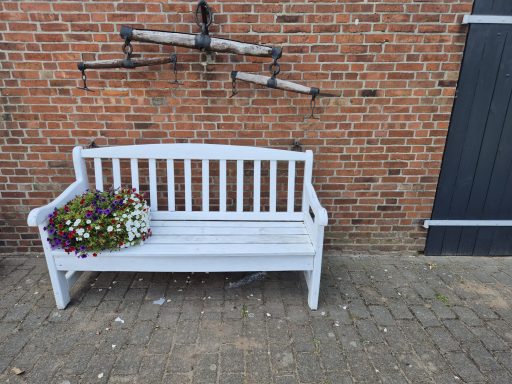 Weiße Bank mit buntem Blumengebinde vor einer roten Backsteinmauer.
