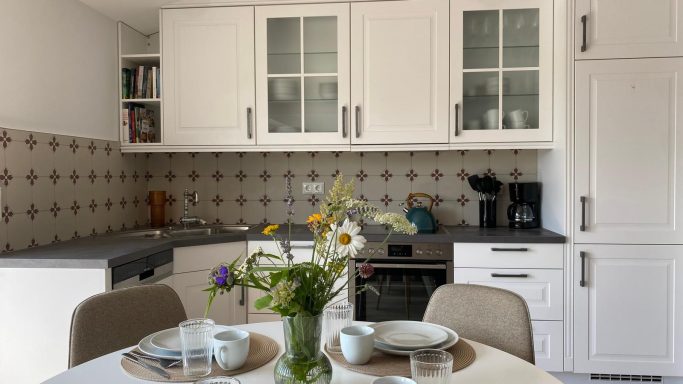 Helle Küche mit weißem Mobiliar, Tisch gedeckt mit Blumen und Geschirr.